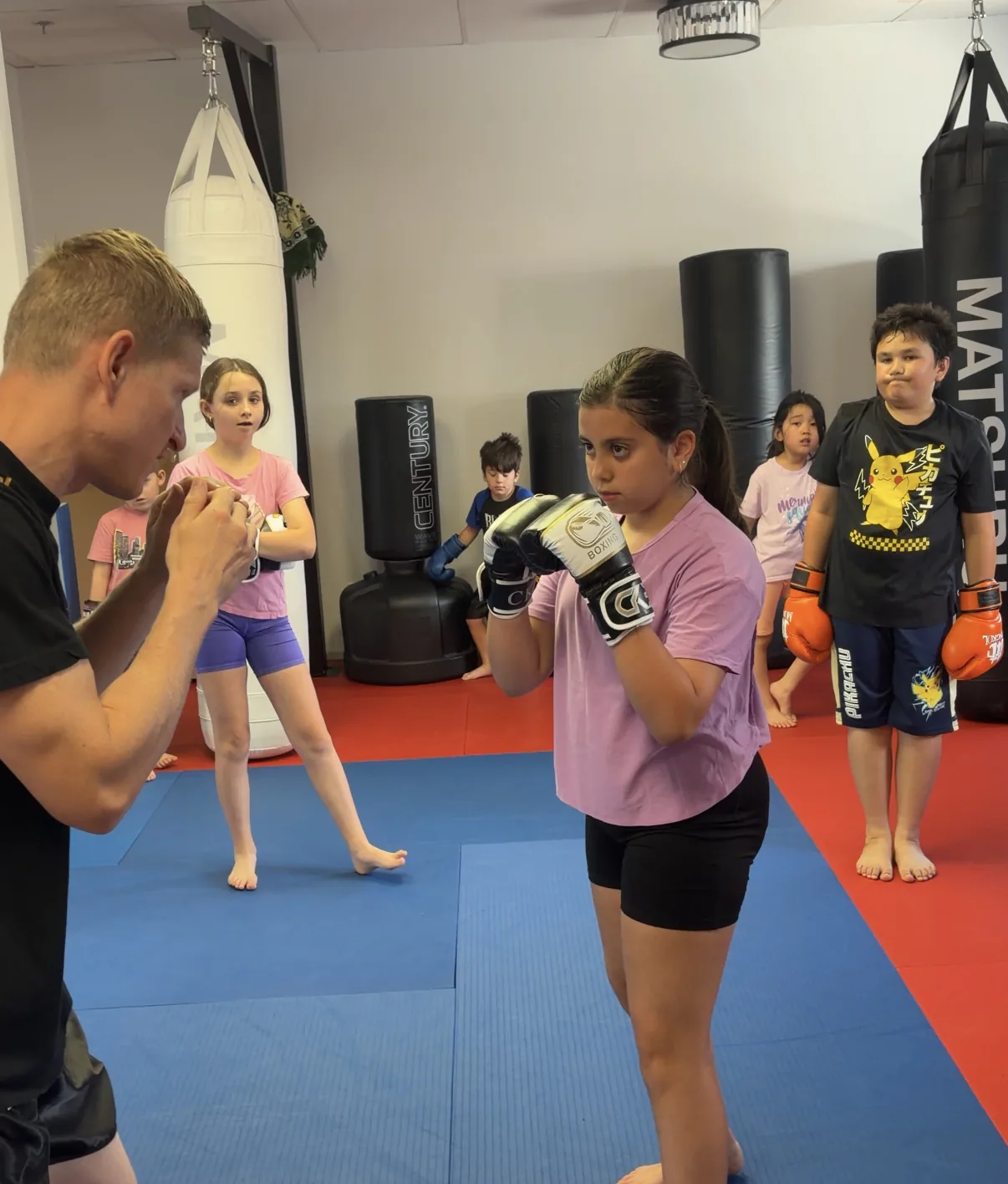 A coach holding focus mitts for a young student during a kids Muay Thai class at Maple Jiu-Jitsu Academy